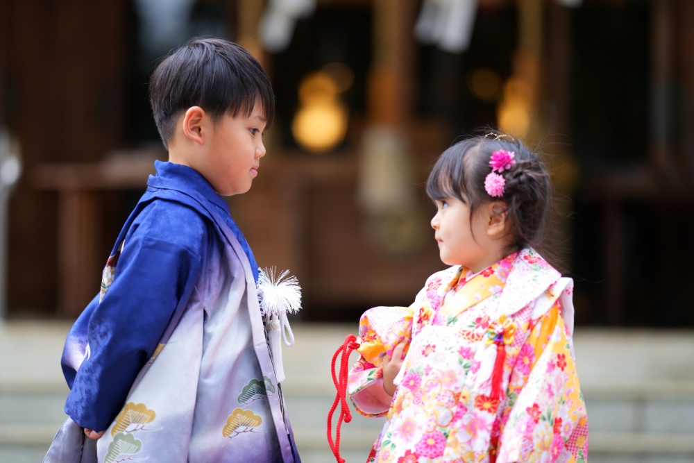 青い着物を着た男の子と、ピンク色の花柄の着物を着た女の子が、寺院の前で向かい合って立っている姿。