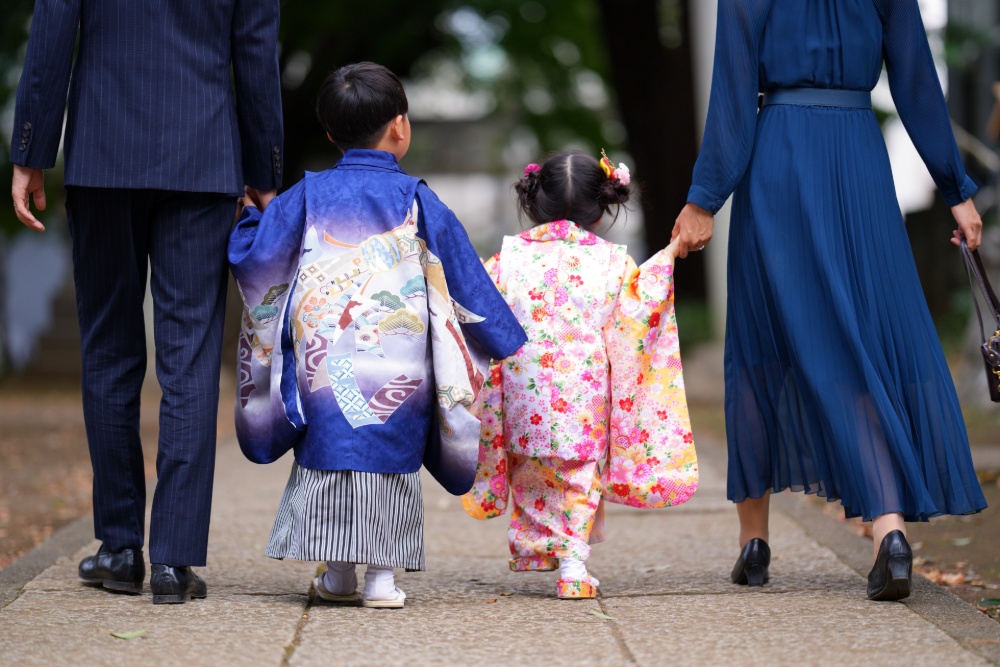 紺色の洋装を着た両親と、青い羽織と白い帯、ピンクの花柄の着物を着た2人の子どもが手をつないで歩いている